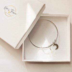 Può includere: Un braccialetto rigido in argento con un ciondolo a forma di luna crescente e una stella. Il braccialetto è in una scatola bianca con il testo "Nala's Treasure Boutique" sul coperchio.