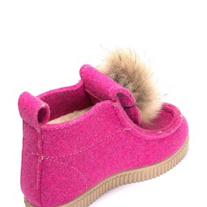 May include: Pink felt boots with a brown fur pom-pom on top. The boots have a beige rubber sole.