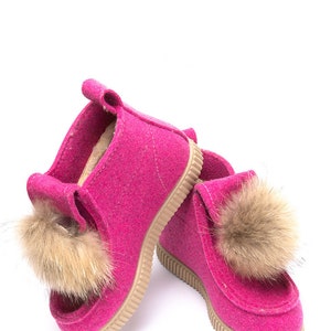 May include: A pair of pink felt boots with brown fur pom-poms. The boots have a rubber sole.