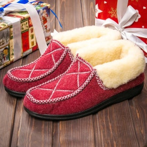 Puede incluir: Un par de zapatillas rojas acolchadas con forro de piel sintética blanca. Las zapatillas tienen un patrón de diamante y están sobre una superficie de madera.