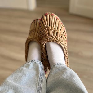 May include: Close-up of woven straw slippers with a braided border and red stitching. The slippers are worn with white socks and light blue denim jeans. The natural colour of the straw contrasts with the white and blue.