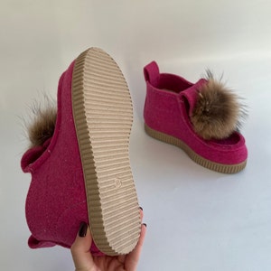 May include: A pair of pink felt slippers with brown fur pom-poms. The slippers have a brown rubber sole with a textured tread.