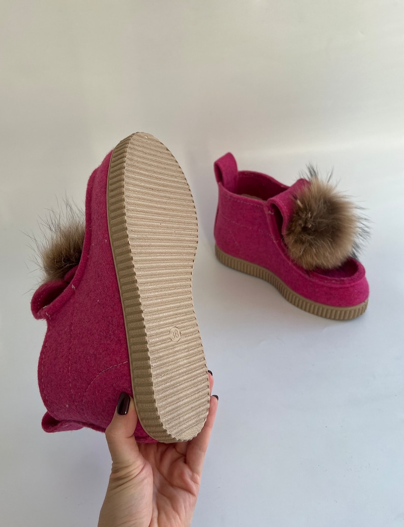 May include: A pair of pink felt slippers with brown fur pom-poms. The slippers have a brown rubber sole with a textured tread.