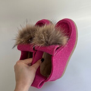 May include: Pink felt slippers with a brown fur pom-pom on top. The slippers have a brown leather sole.