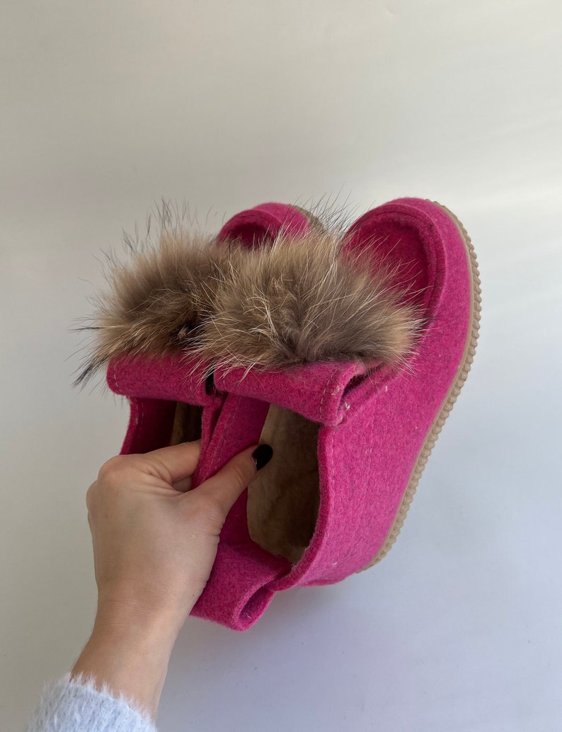 May include: Pink felt slippers with a brown fur pom-pom on top. The slippers have a brown leather sole.