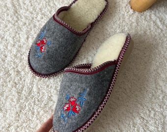 May include: A pair of gray felt slippers with embroidered floral designs in red and blue. The slippers have a cream-colored interior and a dark red trim. The edges are stitched with white thread.
