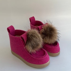 May include: A pair of pink felt boots with brown fur pom-poms. The boots have a rubber sole and a pink felt upper.