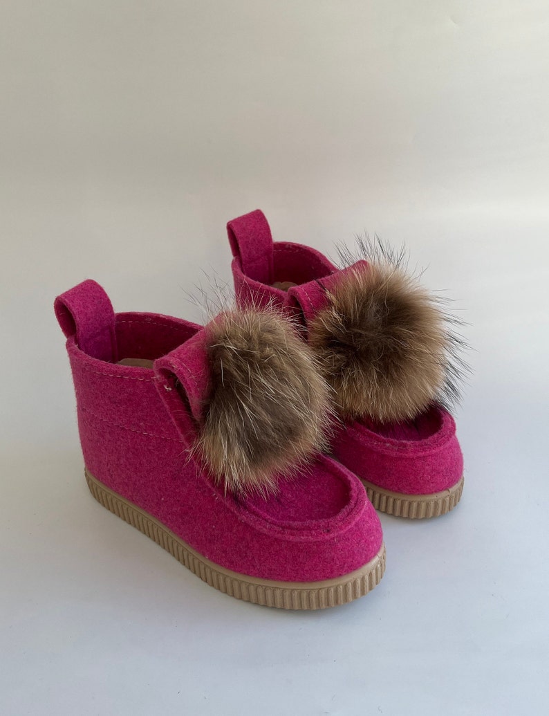 May include: A pair of pink felt boots with brown fur pom-poms. The boots have a rubber sole and a pink felt upper.