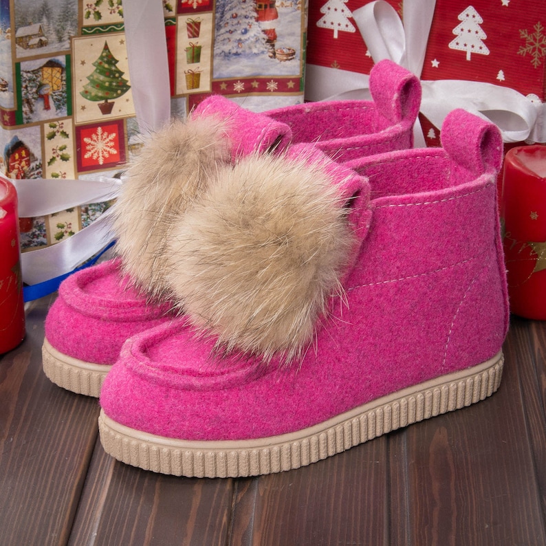 May include: A pair of pink felt boots with faux fur pom-poms. The boots have a white rubber sole and are sitting on a wooden surface.