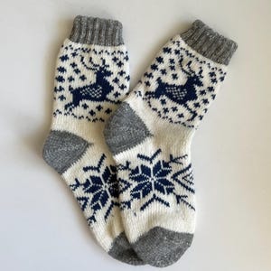 Puede incluir: Un par de calcetines blancos con un puño gris y un patrón de copo de nieve y reno azul y blanco.