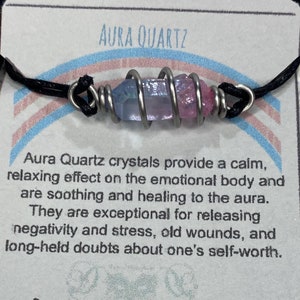 Peut inclure: Un bracelet en cordon noir avec un fil d'argent enroulé autour d'un cristal de quartz aura rose et bleu. La carte attachée au bracelet indique "Aura Quartz" et décrit les propriétés curatives du cristal. La carte comprend également le texte "Denique Vintage Designs" et "Handmade in the Adirondacks".