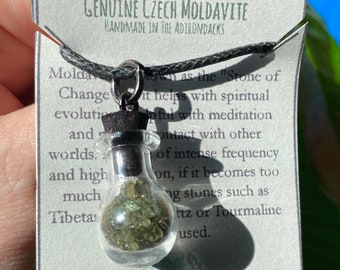 Op de afbeelding: Een glazen flesje-hanger gevuld met groene Moldaviet stenen, hangend aan een zwart koord. Het label leest "GENUINE CZECH MOLDAVITE, HANDMADE IN THE ADIRONDACKS" en "DENIQUE VINTAGE DESIGNS".