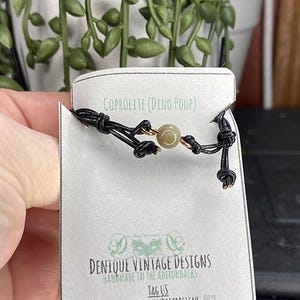 May include: A black cord bracelet with a central beige bead and gold-colored accents. The bracelet is attached to a white tag with the text "COPROLITE (DINO POOP)" and "DENIQUE VINTAGE DESIGNS". The tag also includes the text "HANDMADE IN THE ADIRONDACKS" and a social media handle.