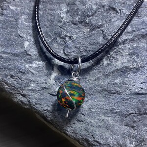 May include: A spherical pendant featuring a dark base with iridescent orange and green flecks, wrapped in silver-toned wire. The pendant hangs from a black cord necklace, secured by a silver-toned jump ring at the top of the wire wrapping.