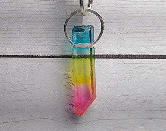 May include: A colorful crystal pendant necklace. The pendant is a rectangular prism with a gradient of colors, including blue, yellow, and pink. It hangs from a silver ring and a black cord. The background is a white wooden surface.