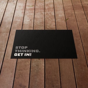 Op de afbeelding: Een rechthoekige zwarte deurmat met zilverkleurige tekst "STOP THINKING. GET IN!". De mat heeft een getextureerd oppervlak en ligt op een houten plankenvloer.