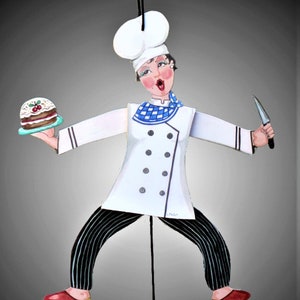 May include: A white chef figurine with a blue and white checkered scarf, holding a cake and a knife. The figurine is wearing a white chef's hat and black and white striped pants.