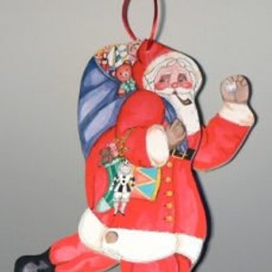 May include: A festive Santa Claus ornament, painted in bright red and white, with a full beard and a sack of toys. The ornament has a red ribbon for hanging and is detailed with a drum and a toy soldier. The Santa is depicted in a running pose.