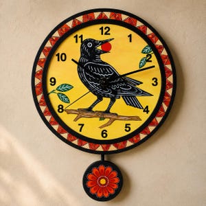 May include: A round wall clock with a yellow face featuring a black crow perched on a branch holding a red berry. The clock has black numbers, black hands, and a decorative border of red and white triangles. A round pendulum with a red flower hangs below.
