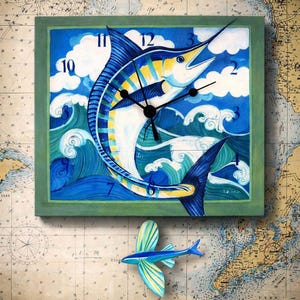 May include: Square wall clock with a painted marlin design in shades of blue, yellow, and white, set against a backdrop of ocean waves and clouds. The clock has black hands and a green frame. A small, blue, decorative flying fish is below the clock.
