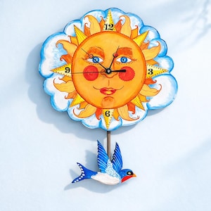 May include: A whimsical wall clock featuring a smiling sun face with blue eyes, red cheeks, and a yellow sunburst design. The clock is surrounded by a blue cloud border and has a pendulum in the shape of a blue and white bird.