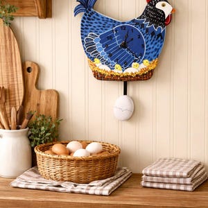 May include: A blue and black chicken-shaped wall clock with a pendulum egg. The clock features a blue face with black hands and a decorative nest with chicks. A woven basket of eggs and folded checkered towels are on the wooden countertop.