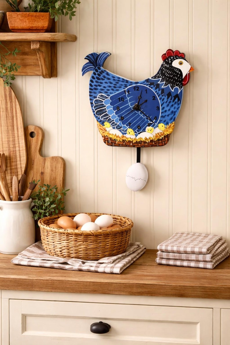 May include: A blue and black chicken-shaped wall clock with a pendulum egg. The clock features a blue face with black hands and a decorative nest with chicks. A woven basket of eggs and folded checkered towels are on the wooden countertop.