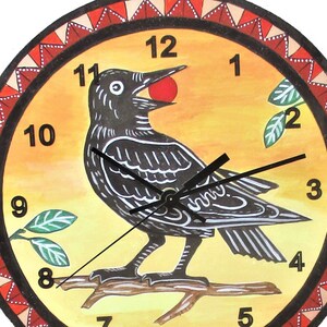 May include: A round wall clock with a yellow and orange gradient background featuring a black bird perched on a branch holding a red berry in its beak. The clock face has black numbers from 1 to 12 and a decorative red and white triangular border.