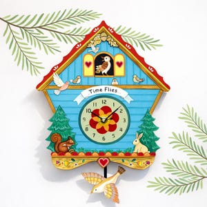 May include: A whimsical cuckoo clock with a blue facade, red roof, and gold trim. The clock face features a floral design and the words "Time Flies." Decorative elements include birds, a squirrel, a rabbit, and a swinging bird pendulum.