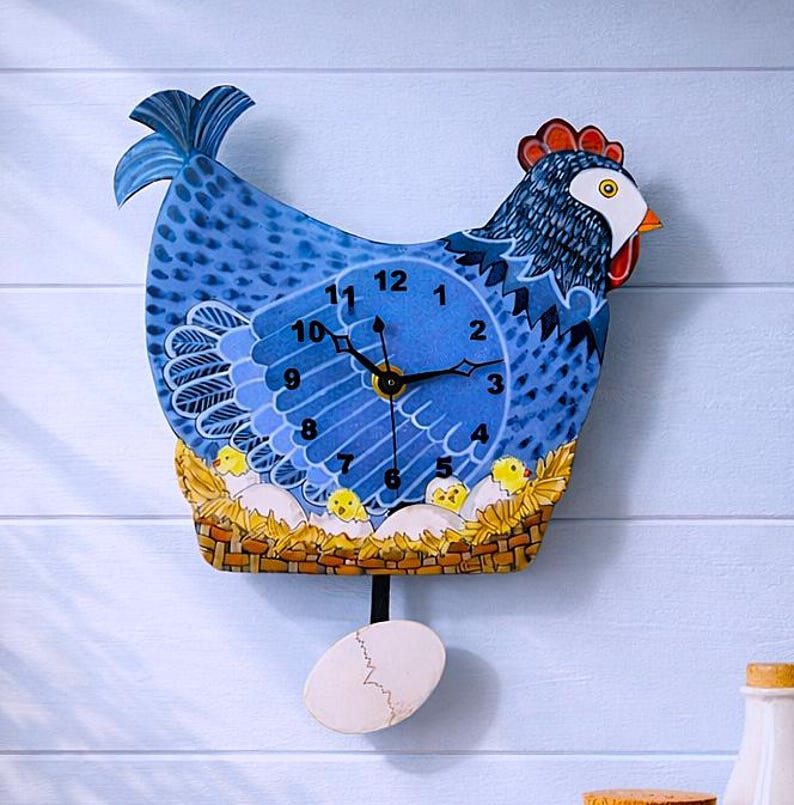 May include: A whimsical blue chicken-shaped wall clock with a pendulum. The clock face features black hands and white numbers. The design includes a nest with chicks and an egg-shaped pendulum. The clock is mounted on a light blue wooden wall.