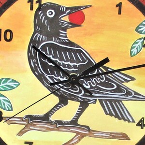 May include: A round wall clock featuring a hand-painted illustration of a black bird perched on a brown branch against a yellow background. The bird holds a small red sphere in its beak. Black clock hands are centered over the bird, with black numbers 1, 3, 4, 7, 8, 10, and 11 visible around the perimeter.