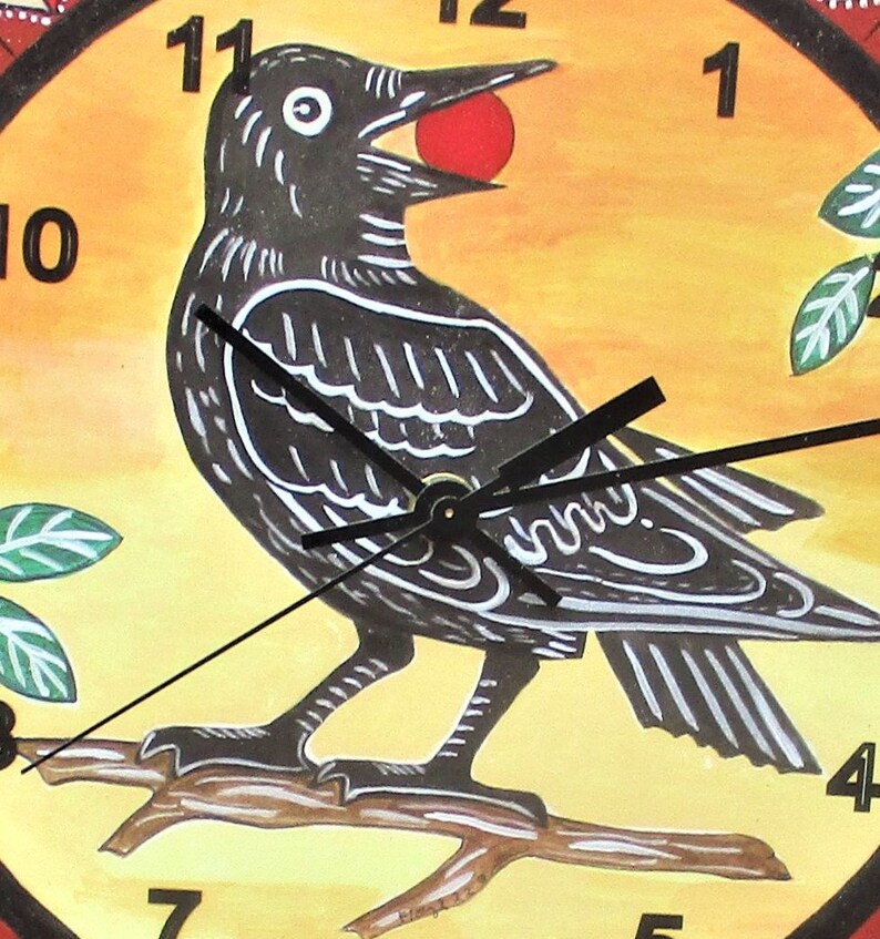 May include: A round wall clock featuring a hand-painted illustration of a black bird perched on a brown branch against a yellow background. The bird holds a small red sphere in its beak. Black clock hands are centered over the bird, with black numbers 1, 3, 4, 7, 8, 10, and 11 visible around the perimeter.