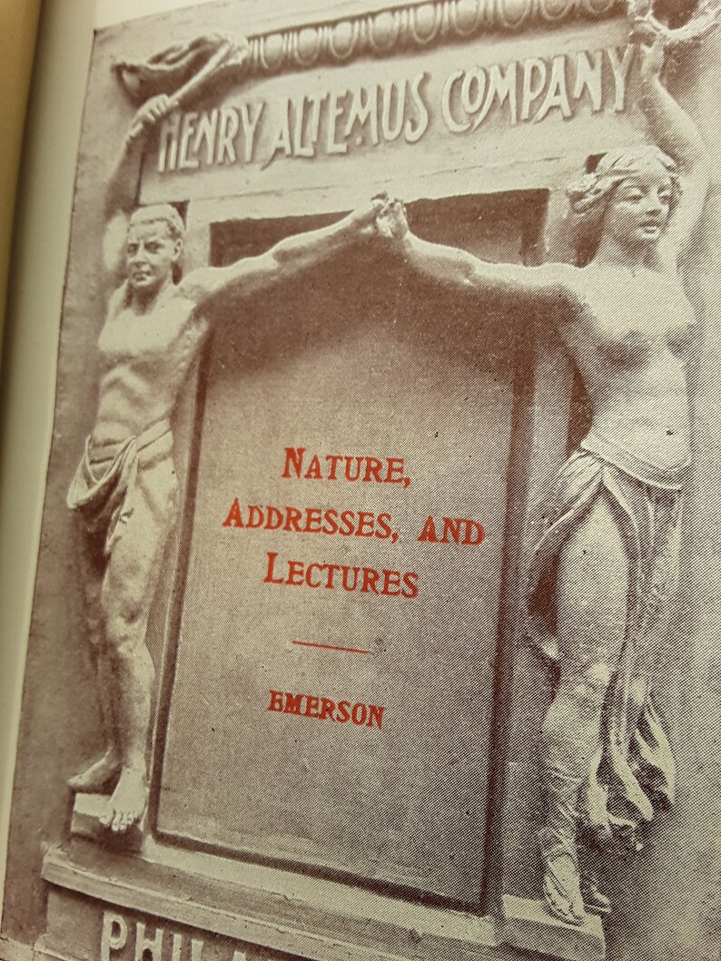 May include: A vintage book cover with a black and white image of two figures holding up a banner that reads "Henry Altemus Company". The text "Nature, Addresses, and Lectures" is printed in red below the banner. The author's name, "Emerson", is printed in red below the text.
