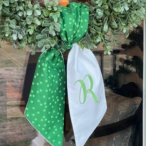 Op de afbeelding: Een groene krans met een decoratieve strik. De strik heeft een groene stof met kleine klaverpatronen en een witte stof met een groen geborduurde letter "R". De krans is gemaakt van groene bladeren.