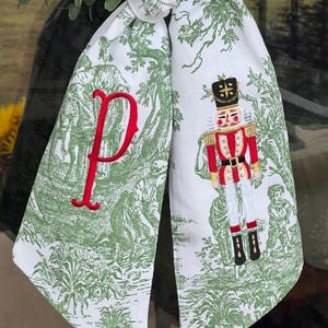 May include: White fabric Christmas decoration with a green toile pattern and a red embroidered letter "P". The decoration features an embroidered nutcracker soldier in a red and gold uniform. The fabric is tied in a knot.