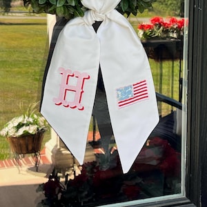 Op de afbeelding: Een witte stoffen kranssjerp met een roze geborduurde letter H aan de linkerkant en een kleine geborduurde Amerikaanse vlag aan de rechterkant. De sjerp is aan de bovenkant geknoopt en hangt met twee puntige uiteinden over een groene krans.
