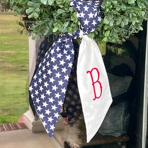 Op de afbeelding: Een decoratief kransaccessoire met een marineblauwe stoffen staart met witte sterren en een witte stoffen staart met een rode geborduurde letter B. De twee stoffen delen zijn aan elkaar geknoopt en bevestigd aan een groene bladerkrans.