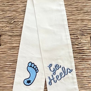 Op de afbeelding: Witte stoffen sjerp met blauwe borduursels van een voetafdruk en de tekst "Go Heels".
