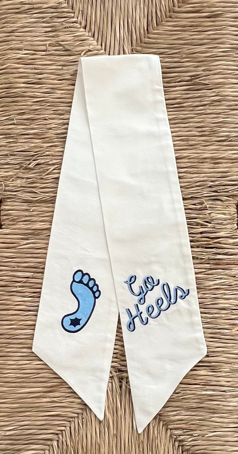 Op de afbeelding: Witte stoffen sjerp met blauwe borduursels van een voetafdruk en de tekst "Go Heels".