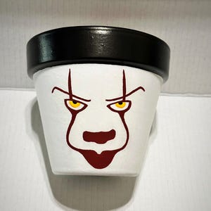 Może przedstawiać: Biała ceramiczna doniczka z ręcznie malowanym wzorem twarzy klauna. Doniczka posiada grubą czarną krawędź u góry. Twarz klauna obejmuje żółte oczy z ciemnoczerwonymi konturami, czerwone brwi oraz czerwoną nos i usta namalowane na białej powierzchni.