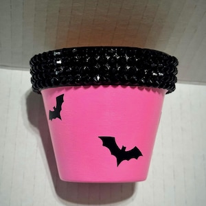 May include: A bright pink plant pot featuring black bat silhouettes scattered across the exterior. The upper rim of the pot is decorated with a thick band of faceted black beads that create a textured, sparkling border around the top edge.