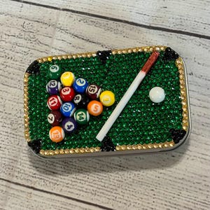 May include: A rectangular metal tin decorated with green, gold, and black rhinestones arranged to resemble a pool table. The surface features a miniature rack of numbered billiard balls, a white cue ball, and a small pool cue stick.