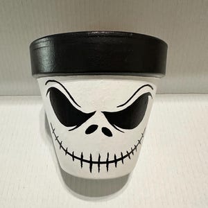 May include: A white ceramic plant pot featuring a hand-painted black rim and a stylized face design. The face consists of two large, almond-shaped black eyes and a wide, stitched-style mouth line painted in black across the lower section.