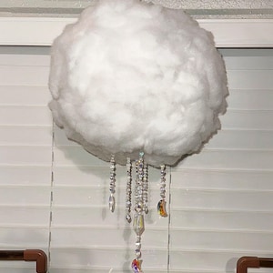 Puede incluir: Una decoración colgante con forma de nube blanca y esponjosa hecha de material suave. Varios hilos de cuentas de cristal transparente facetadas cuelgan de la parte inferior, con colgantes iridiscentes en forma de luna y prismas de vidrio.