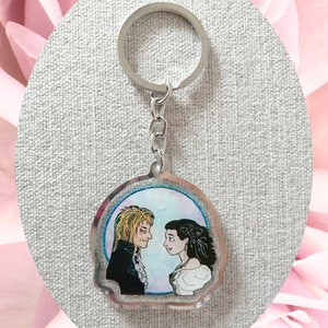 May include: A silver keychain with a clear round charm featuring a hand-drawn illustration of two people looking at each other. The illustration is set against a blue and pink watercolor background.