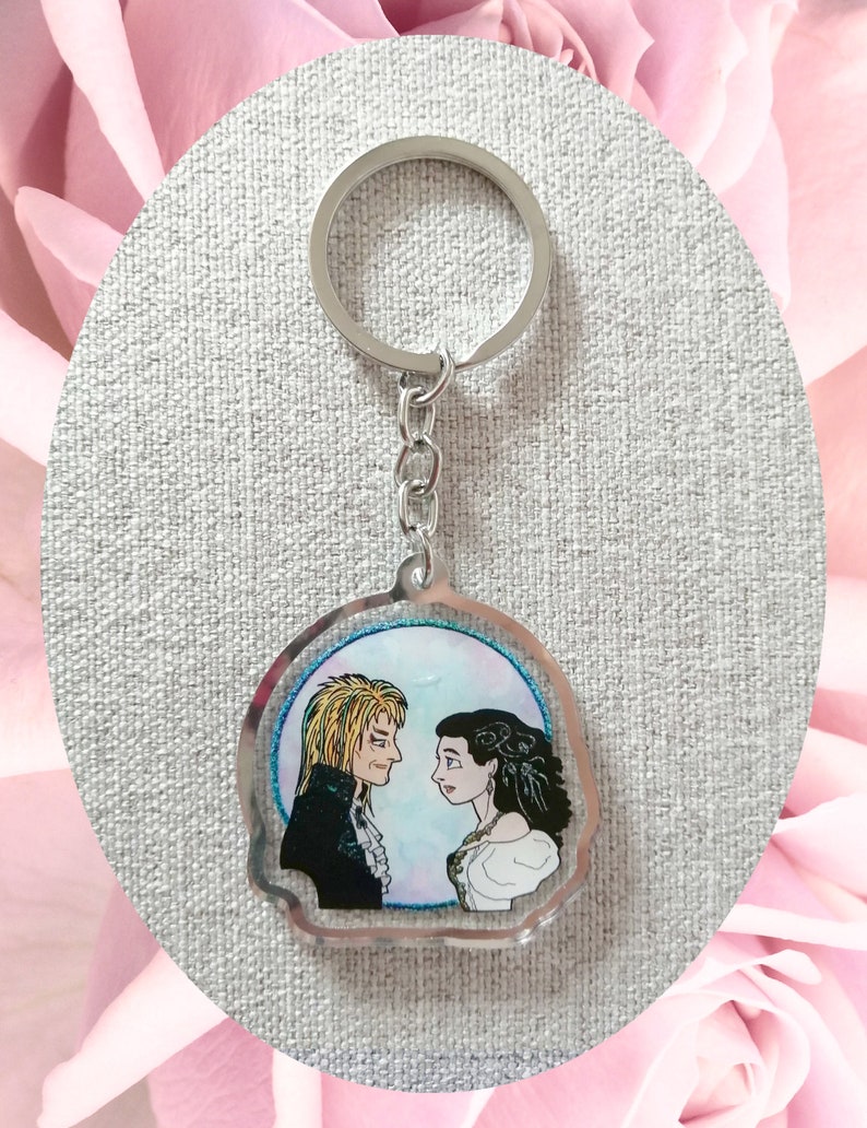 May include: A silver keychain with a clear round charm featuring a hand-drawn illustration of two people looking at each other. The illustration is set against a blue and pink watercolor background.