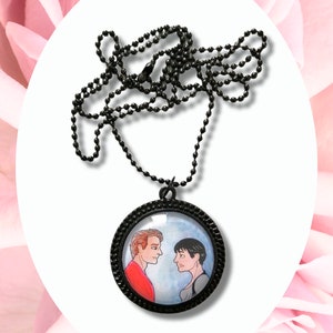May include: A black chain necklace with a round glass pendant featuring a hand-painted illustration of two people looking at each other. The pendant has a black metal frame.