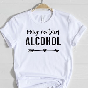 May include: White t-shirt with the text "may contain ALCOHOL" in black, with an arrow and heart design. The shirt is hanging on a wooden hanger against a white wooden background. The t-shirt is made of a soft material.