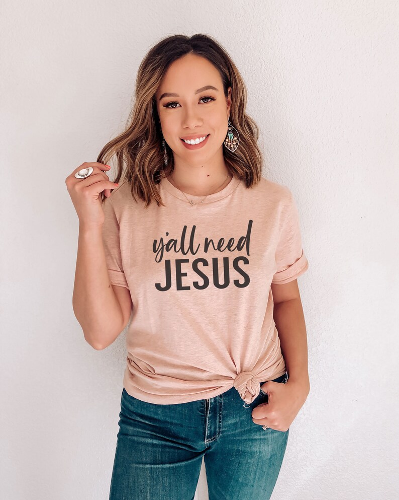 May include: A woman wearing a light pink t-shirt with the text "y'all need JESUS" in black lettering. She is wearing blue jeans and has her hair pulled back.