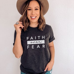 May include: A woman wearing a dark gray t-shirt with the text "FAITH over FEAR" in white. She is also wearing a brown hat and blue jeans.